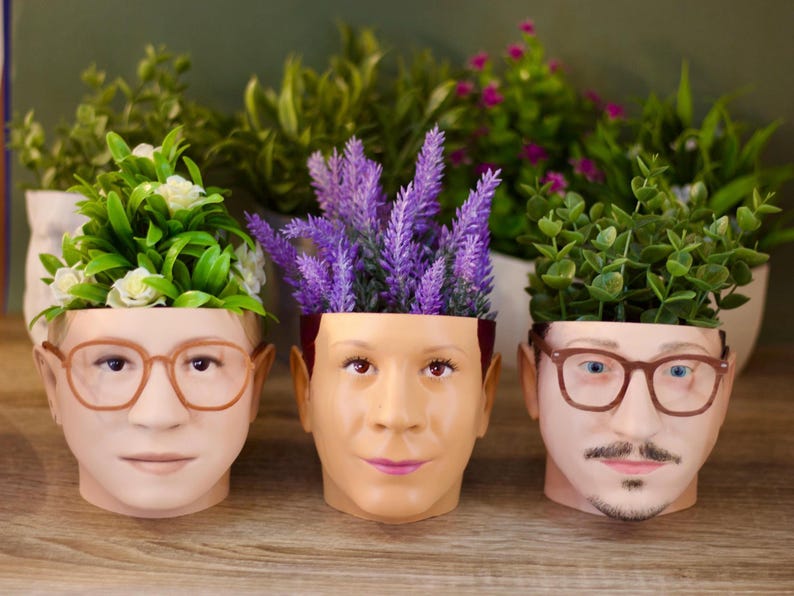 May include: Three ceramic planters shaped like human faces, each with a different plant growing out of the top. The planter on the left has a green plant with white flowers, the middle planter has purple flowers, and the planter on the right has a green plant.
