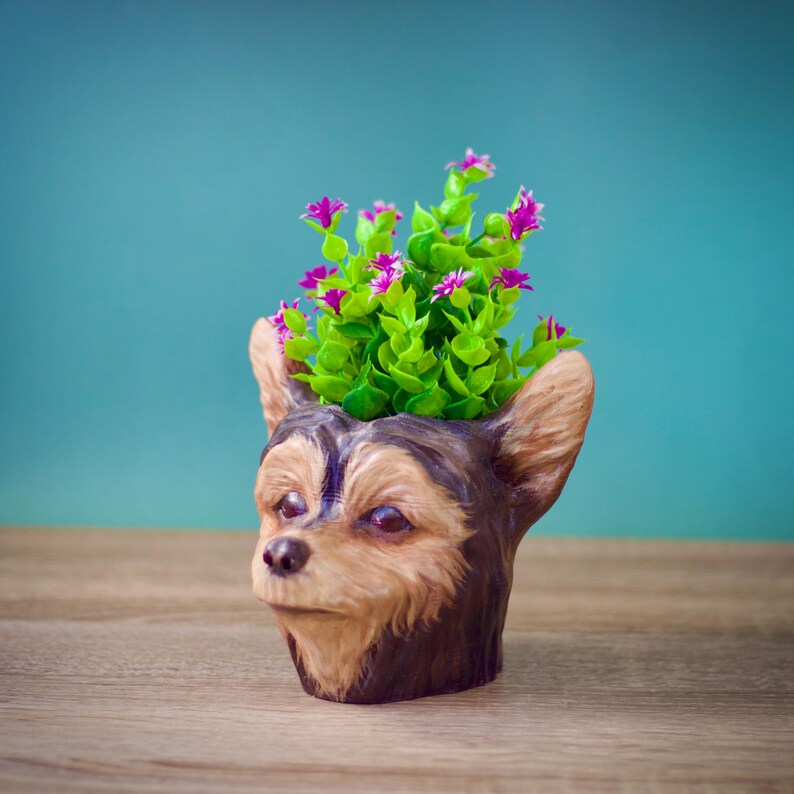 May include: A brown and black ceramic planter shaped like a Yorkshire Terrier dog's head with a green plant with pink flowers growing out of the top.