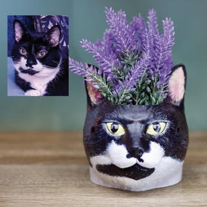 May include: A black and white cat planter with a purple flower arrangement. The planter is shaped like a cat's head with a white face and black ears.