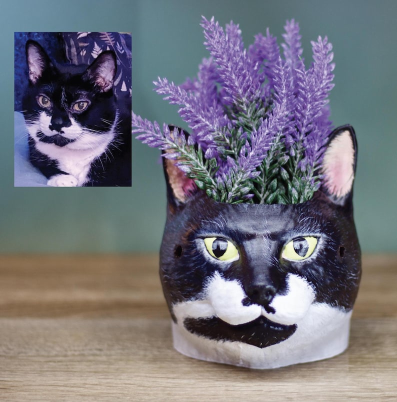 May include: A black and white cat planter with a purple flower arrangement. The planter is shaped like a cat's head with a white face and black ears.