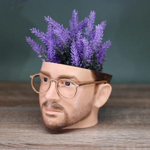 May include: A ceramic planter shaped like a person's head with glasses, filled with purple lavender flowers.