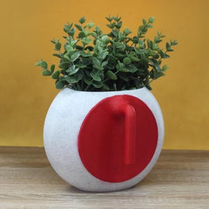 May include: A white speckled ceramic planter with a large red circle and handle detail. The planter is filled with green leafy faux plants. The background is a solid yellow color.