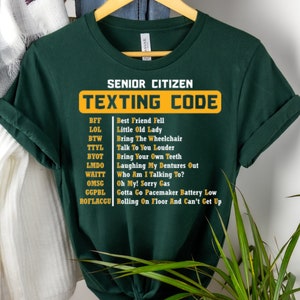 Op de afbeelding: Een groen T-shirt met de tekst "Senior Citizen Texting Code" in geel. Het shirt geeft een lijst met veelgebruikte sms-afkortingen voor senioren en hun betekenis.