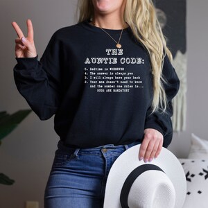 Peut inclure: Un sweat-shirt noir avec du texte blanc qui dit "The Auntie Code:" et énumère cinq règles, dont "Bedtime is whenever" et "Hugs are mandatory".
