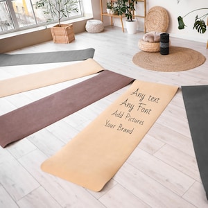 May include: Five rectangular yoga mats in shades of charcoal gray, beige, and brown are laid out on a light wood floor. One beige mat features black text that reads: Any text, Any Font, Add Pictures, Your Brand, in a simple handwritten style.