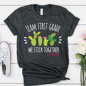 以下が含まれることがあります： 濃いグレーのTシャツに「Team First Grade We Stick Together」という文字と、ピンクの花が咲いたサボテン4本のグラフィックがプリントされています。「Miss Reinke」という文字がサボテンの下にあります。