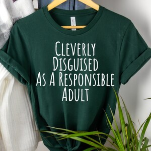 Könnte beinhalten: Ein dunkelgrünes T-Shirt mit weißem Text, der "Cleverly Disguised as a Responsible Adult" lautet.
