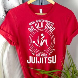 May include: A red t-shirt with white text and a graphic. The text reads "NEVER UNDERESTIMATE AN OLD MAN WHO KNOWS JUIJITSU" around a circular design with two stylized figures. The shirt is displayed on a wooden hanger.