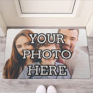 Può includere: Un tappetino bianco per la porta con il testo "YOUR PHOTO HERE" in lettere bianche e nere. Il tappetino presenta una famiglia di tre persone, una donna, un uomo e un bambino, che sorridono alla telecamera.