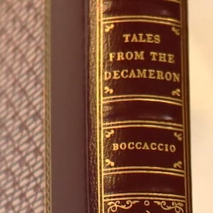 May include: Close-up of a book spine with the title "Tales From The Decameron" by Boccaccio. The dark burgundy cover features gold lettering and decorative accents. The book is a collector's edition.