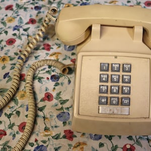 May include: A vintage cream-colored telephone with a push-button keypad. The phone has a coiled cord and a handset. The keypad features numbers and symbols. The phone is set against a floral patterned background.