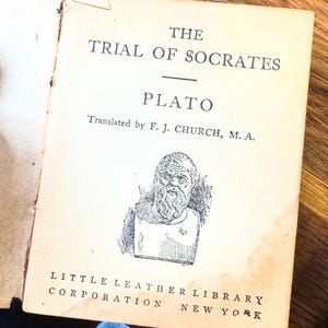 May include: A vintage book cover with a black and white illustration of Socrates. The title is "The Trial of Socrates" by Plato, translated by F. J. Church, M.A. The book is published by Little Leather Library Corporation, New York.