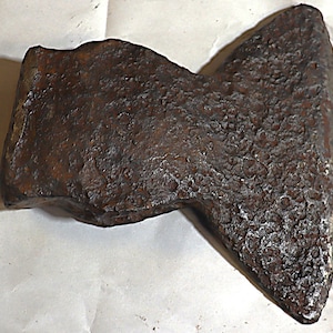 May include: A weathered, antique axe head with a dark brown, textured surface. The axe head has a wide blade and a slightly curved top, suggesting it was used for chopping or splitting wood. The metal shows signs of age and wear.