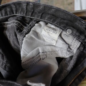May include: Close-up of the inside of a pair of black denim jeans. The jeans have a zipper fly and a label that reads "R1 65-010 Cotton".