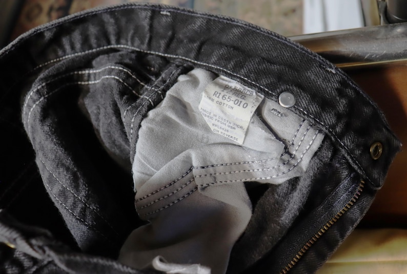 May include: Close-up of the inside of a pair of black denim jeans. The jeans have a zipper fly and a label that reads "R1 65-010 Cotton".
