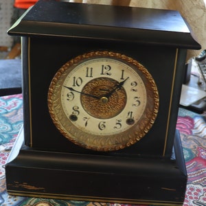 May include: A black wooden mantel clock with a gold-toned face and Roman numeral hour markers. The clock has a decorative gold-toned design around the face and a black pendulum.