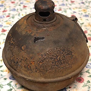 May include: A vintage, rusted, spherical metal Toledo Torch with a top burner assembly. The embossed text reads: THE TOLEDO TORCH, THE TOLEDO PRESSED STEEL CO, TOLEDO OHIO USA, PAT 1702708. The surface shows heavy oxidation and dark brown patina.