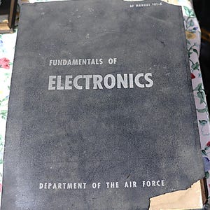 May include: A vintage, dark gray book titled "Fundamentals of Electronics" with the text in silver. The cover also includes "Department of the Air Force" and "AF Manual 101-6". The book shows signs of wear.
