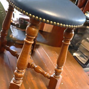 Op de afbeelding: Een vintage houten kruk met een ronde, marineblauwe leren zitting, ongeveer 30 cm in diameter. De zitting is afgewerkt met messingkleurige studs. De gedraaide houten poten zijn verbonden door een onderste spijl. Een klassiek meubelstuk.