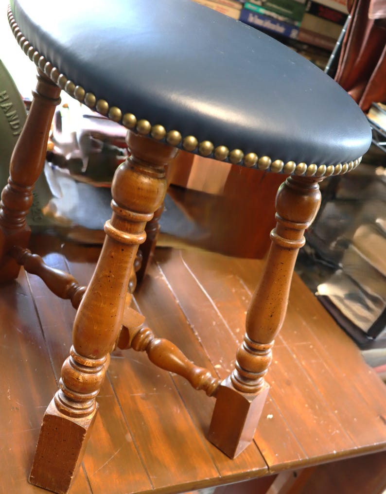 Op de afbeelding: Een vintage houten kruk met een ronde, marineblauwe leren zitting, ongeveer 30 cm in diameter. De zitting is afgewerkt met messingkleurige studs. De gedraaide houten poten zijn verbonden door een onderste spijl. Een klassiek meubelstuk.