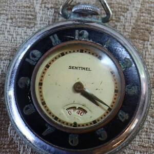 May include: A silver pocket watch with a black face and white numerals. The watch has a small second hand and a 'SENTINEL' brand name.
