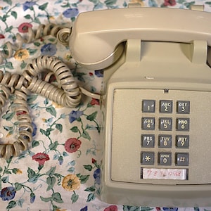 May include: A vintage beige push-button telephone with a coiled cord. The phone has a keypad with numbers and letters, and a sticker with a phone number. The phone is on a floral patterned fabric.