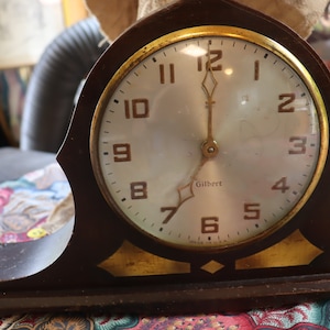 May include: A vintage wooden mantle clock with a white face and gold accents. The clock face reads "Gilbert" and has black Roman numeral hour markers. The clock is set to 12:00.
