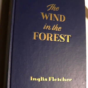 May include: A hardcover book with a dark blue cover. The title "The Wind in the Forest" and author "Inglis Fletcher" are printed in gold lettering. The book's spine is visible.