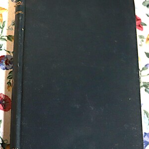 May include: A vintage hardcover book with a dark teal cover. The spine has gold lettering and detailing. The book is slightly worn, suggesting age and use. The book is resting on a floral patterned surface.