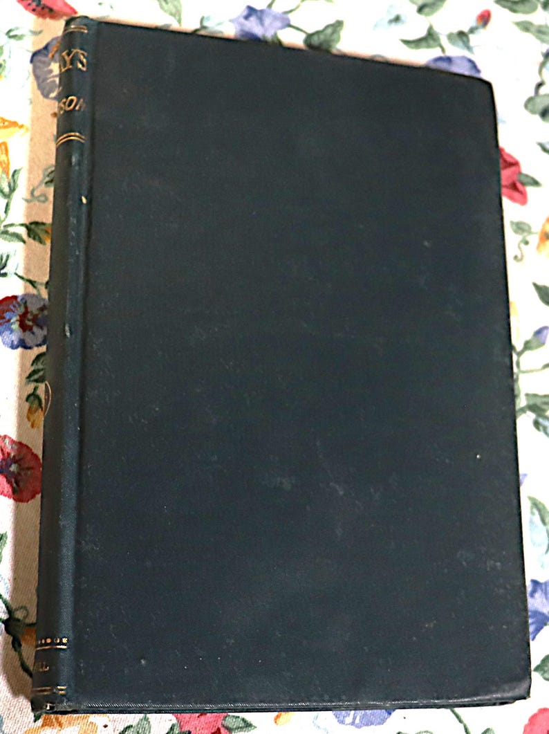 May include: A vintage hardcover book with a dark teal cover. The spine has gold lettering and detailing. The book is slightly worn, suggesting age and use. The book is resting on a floral patterned surface.