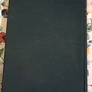 May include: A dark green book with a textured cover. The spine is visible on the right side, with gold accents. The book is resting on a floral patterned surface.