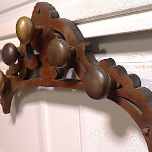 May include: A wall-mounted coat rack featuring a dark brown wooden arched frame with decorative scalloped edges. The rack includes five round, metallic, brass-toned knobs attached to the front, secured with visible screws at the ends.