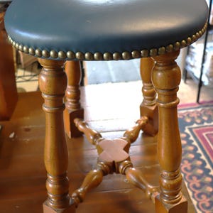 Op de afbeelding: Een vintage houten kruk met een ronde, blauwgroene zitting. De zitting is afgezet met messingkleurige studs. De kruk heeft vier gedraaide poten en een kruisvormige steunstructuur, allemaal in een warme houttint.