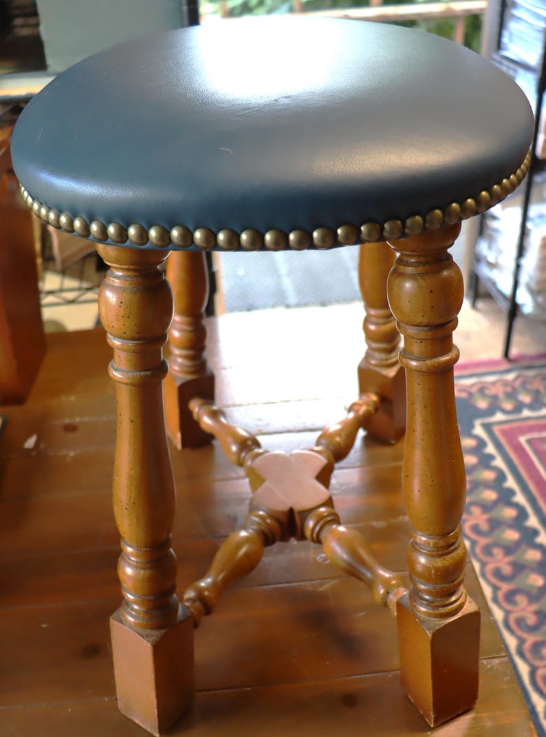 Op de afbeelding: Een vintage houten kruk met een ronde, blauwgroene zitting. De zitting is afgezet met messingkleurige studs. De kruk heeft vier gedraaide poten en een kruisvormige steunstructuur, allemaal in een warme houttint.