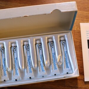 May include: A white box with a green trim containing seven small tubes of white toothpaste labeled "Thermodent" in blue lettering. The box is open and the tubes are visible. A white pamphlet with a blue border and the text "Thermodent Tooth Paste" is visible in the background.