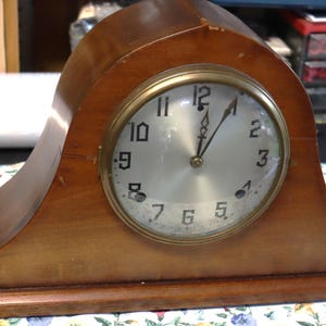 May include: A wooden mantel clock with a light brown finish. The clock face is round with a gold-colored bezel and a silver-colored face. The clock hands point to approximately 12:00. The clock is sitting on a floral patterned surface.