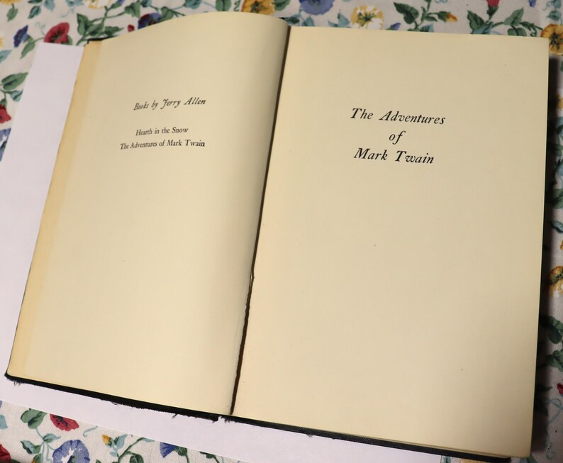 May include: An open book with cream-colored pages. The left page displays the text "Books by Jerry Allen", followed by "Hearth in the Snow" and "The Adventures of Mark Twain". The right page reads "The Adventures of Mark Twain".