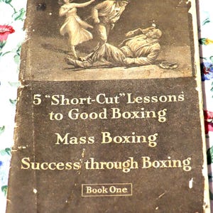 May include: Vintage book cover with sepia-toned illustration of a boxing scene. The cover reads "5 'Short-Cut' Lessons to Good Boxing" and "Success through Boxing." Published by Marshall Stillman Association, New York. Book One.