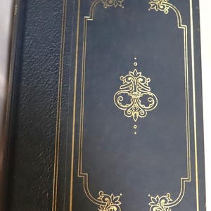May include: A hardcover book with a dark blue cover and gold decorative accents. The cover features a gold ornate design in the center and gold borders. The spine is a darker blue with a textured appearance.