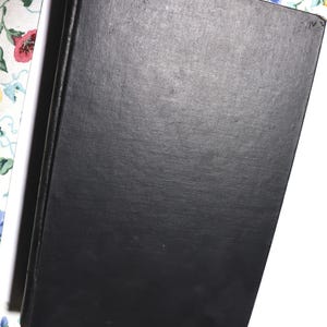 May include: A black hardcover book with a textured cover. The book is slightly angled, revealing a white edge and a floral patterned background.