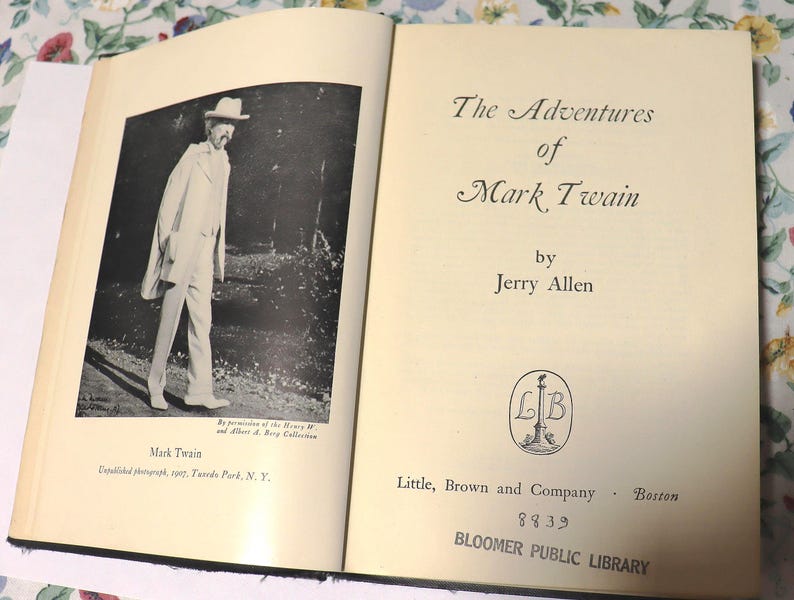 May include: An open book featuring a black and white photograph of Mark Twain and the title "The Adventures of Mark Twain" by Jerry Allen. The book is from the Bloomer Public Library.