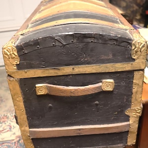 May include: An antique trunk with a curved top, featuring a black exterior with brass accents and a leather handle. The trunk has a wooden frame with metal corner protectors and brass bands. The top is a lighter brown color.