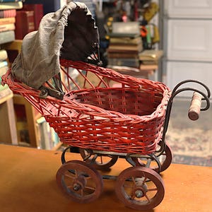 May include: A vintage-style doll carriage featuring a bright red woven wicker body, a dark gray fabric hood, and four brown wooden wheels. The carriage includes a metal frame with a wooden handle grip, showcasing a classic design with visible woven textures.