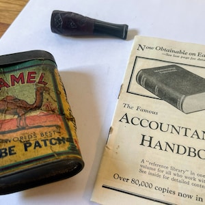 May include: A vintage tin box with a camel illustration and the text "Camel The World's Best Tube Patch".  The box is next to a brown and black pipe and a pamphlet titled "The Famous Accountants' Handbook".