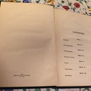 May include: An open book with aged, off-white pages. The right page displays the word "CONTENTS" at the top, followed by a list of essays and page numbers. The book has a dark blue cover and is resting on a floral patterned surface.