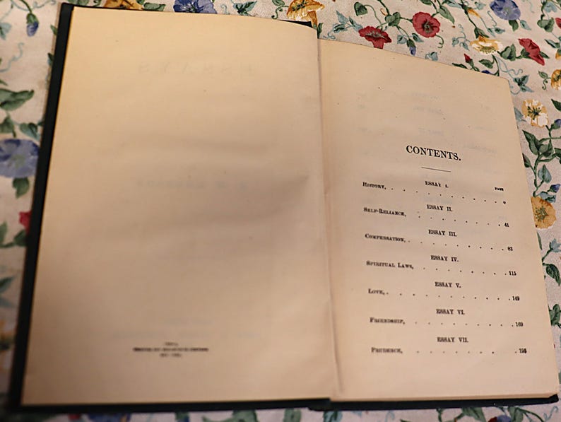 May include: An open book with aged, off-white pages. The right page displays the word "CONTENTS" at the top, followed by a list of essays and page numbers. The book has a dark blue cover and is resting on a floral patterned surface.