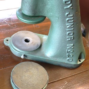 May include: A vintage green metal Tandy Junior No. 20 press machine. The press has a circular base and a lever arm. The machine is on a wooden surface. The press is designed for crafting and leatherwork.