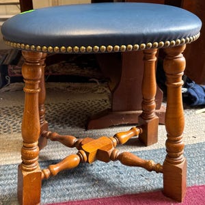 Op de afbeelding: Een kleine, ronde houten kruk met een marineblauwe leren zitting. De zitting is afgewerkt met goudkleurige studs. De poten zijn gedraaid hout en de kruk heeft een kruisvormige sprei voor extra stabiliteit. Een klassiek meubelstuk.