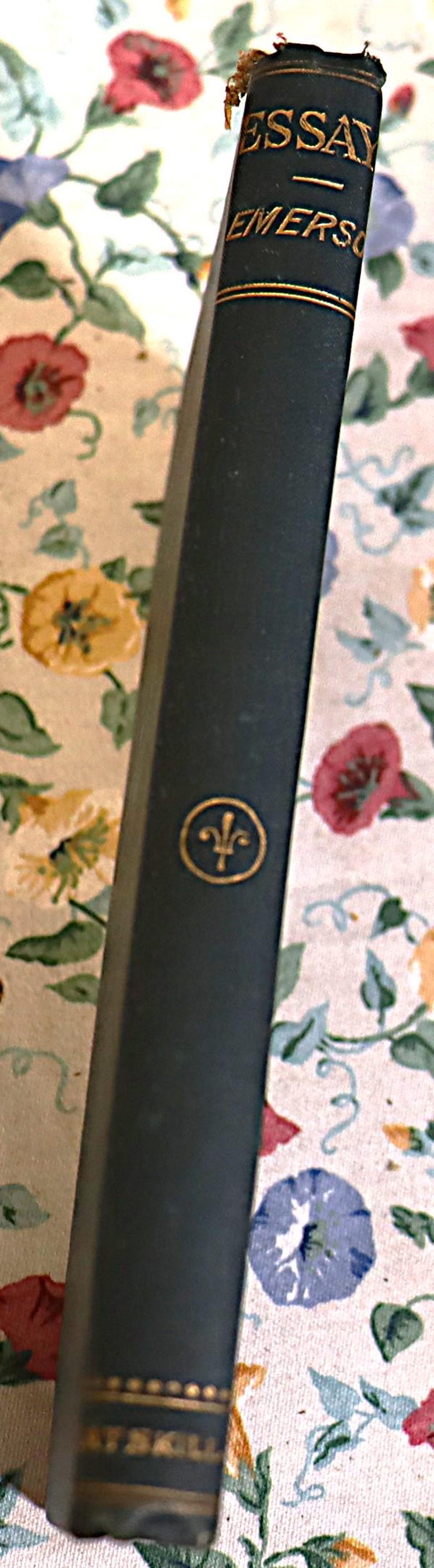 May include: A dark green book with gold lettering that reads "ESSAY EMERSON" on the spine. A gold emblem is centered on the book's cover. The book is set against a floral patterned background.