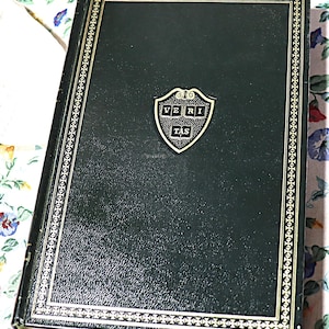 May include: A dark green book with a textured cover and gold-colored trim. A gold-colored emblem with the word "VERITAS" is centered on the cover. The book is resting on a floral patterned surface.